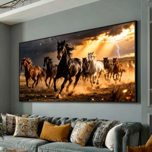 Op de afbeelding: Een ingelijste canvas print met een kudde paarden die over een veld galopperen. De paarden zijn bruin, zwart en wit, tegen een dramatische lucht met bliksem. Het kunstwerk wordt tentoongesteld boven een grijze bank met decoratieve kussens.