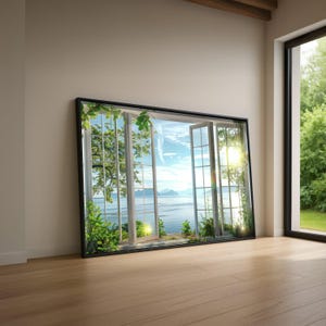 Puede incluir: Una gran obra de arte enmarcada que representa una ventana abierta con vistas a una serena escena costera. El marco negro rodea una vista del cielo azul, el mar y la exuberante vegetación. La obra está apoyada contra una pared sobre un suelo de madera.