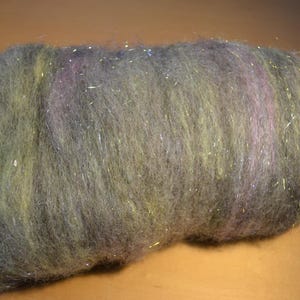 May include: A close-up of a bundle of fiber for crafting. The fiber is a mix of gray, green, and purple with shimmering silver strands throughout. The texture appears soft and fluffy, suitable for various textile projects.