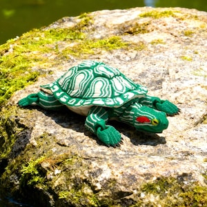 May include: A green and white plush turtle toy with a detailed shell pattern and red accents on its head. The turtle is resting on a moss-covered rock, with a blurred green background suggesting a natural setting.