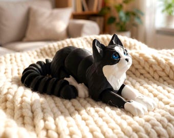 May include: A black and white cat figurine with blue eyes, lying on a cream-colored, chunky knit blanket. The cat has a long, segmented tail and white paws. The figurine is a decorative item.