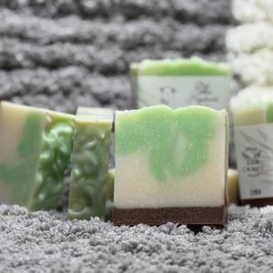 May include: Several handmade soaps with a layered design. The soaps have a light green and white swirl pattern, with a dark brown base. Some soaps have a textured surface. The soaps are on a gray and white textured surface.