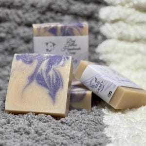 May include: Several bars of handmade soap with a creamy beige base and purple swirls. Each bar has a white label with the text "E&B Craft" and a logo. The soaps are arranged on a gray and white textured surface.