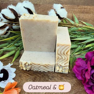 May include: Three bars of oatmeal soap with a light beige color and visible oatmeal flakes. The soap is arranged on a wooden surface with decorative flowers and greenery. The text "Oatmeal &" is visible on a pink label.