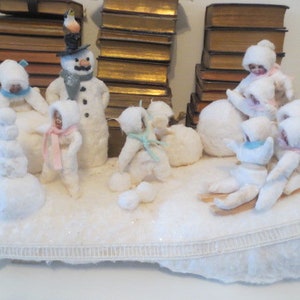 May include: A winter-themed display featuring several small, white snowman figures with various colored scarves and hats. Some snowmen are standing, others are seated or on sleds, all on a white, textured base. A larger snowman with a top hat is also present.
