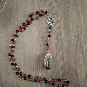 May include: A necklace featuring alternating black and red beads linked by silver-toned metal chain. A silver-toned pentagram charm is positioned near the top, and a single, wire-wrapped, translucent dark-toned crystal point hangs as a pendant.