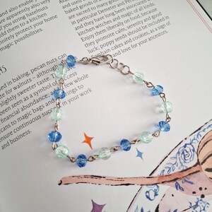 May include: A delicate bracelet featuring alternating light blue and clear beads strung on a silver chain. The bracelet has a lobster clasp closure. The beads are faceted, catching the light. The bracelet is displayed on a printed page.