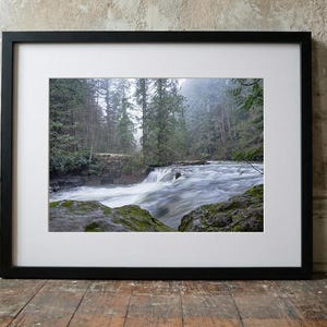 Peut inclure: Photographie encadrée d'une rivière traversant une forêt. L'eau est floue, suggérant le mouvement, et les arbres sont grands et verts. Le cadre est noir, et la photo est montée avec un bord blanc.