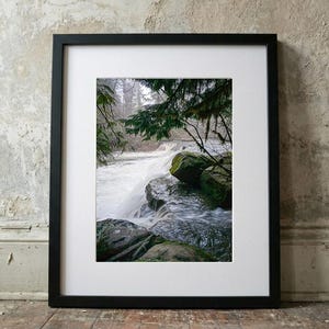 Peut inclure: Photographie encadrée d'une scène de cascade. L'image présente une cascade dévalant des rochers couverts de mousse, encadrée par un feuillage vert. La photographie est dans un cadre noir avec un passe-partout blanc, posée sur une surface en bois.