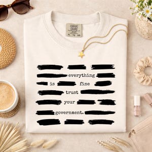 Może przedstawiać: Kremowa koszulka Comfort Colors z napisem "everything is fine trust your government." w kolorze czarnym. Widoczny złoty naszyjnik z gwiazdką i okulary przeciwsłoneczne.