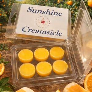 Puede incluir: Un recipiente de plástico transparente que contiene seis ceras aromáticas redondas y amarillas. El recipiente está abierto, revelando las ceras. La etiqueta superior dice "Sunshine Creamsicle" con un logotipo. El fondo muestra un huerto de naranjos.
