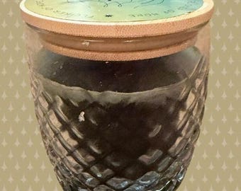 May include: A clear glass jar with a diamond-cut textured pattern filled with dark wax. The jar features a light-colored wooden lid with a circular label on top displaying a blue and green gradient, a line drawing of hands, and the text FLICKER OF HOPE.