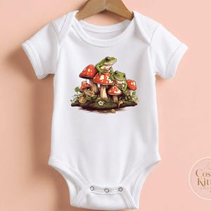 May include: A white short-sleeve baby bodysuit featuring a central graphic of two green frogs perched on a cluster of red and white spotted mushrooms with green foliage. The bottom right corner displays the text Cosmic Kitten Couture.