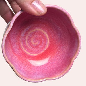 May include: A small, handmade ceramic bowl with a scalloped edge. The bowl is a vibrant pink with a lighter pink and yellow spiral design in the center. The rim has a subtle, natural color.