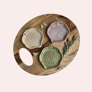 May include: Three ceramic graters in speckled cream, light purple, and sage green, each featuring a scalloped edge and a central textured surface for grating, arranged on a round wooden board with a handle cutout and a sprig of rosemary.
