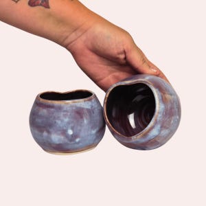 May include: Two small, rounded ceramic cups featuring a glossy glaze in shades of purple, blue, and brown. The vessels have an irregular, organic rim shape and a smooth, reflective surface, showcasing a blend of cool and warm tones throughout.