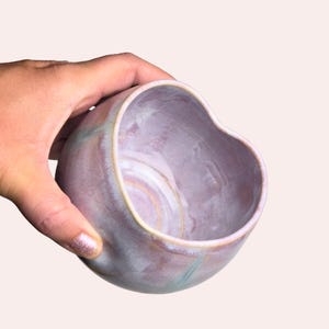 May include: A heart-shaped ceramic bowl with a marbled lavender and blue glaze. The bowl has a rounded shape and a smooth, glossy finish. The bowl is held in a hand.