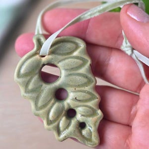 May include: A ceramic leaf pendant in shades of green, with three circular cutouts and a textured surface. The pendant is suspended from a light green ribbon, suggesting it's a necklace or ornament.