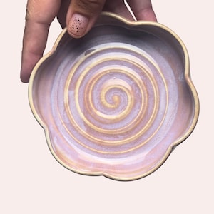 May include: A flower-shaped ceramic dish with a glossy finish, featuring a light purple and soft pink glaze. The interior surface displays a prominent, raised spiral pattern in a creamy beige tone that radiates from the center to the edges.