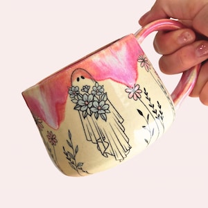 May include: A ceramic mug with a cream-colored base and a glossy pink drip glaze around the rim. The mug features a black line art illustration of a ghost holding a bouquet of flowers, surrounded by additional floral and botanical line drawings.