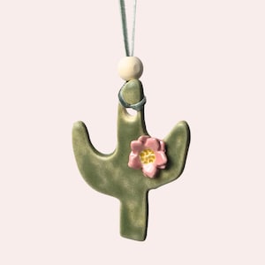 May include: A green ceramic cactus ornament with a pink flower and a light green ribbon for hanging. The ornament has a wooden bead and a silver ring at the top. The cactus is a decorative item.