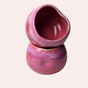 May include: Two stacked, handmade ceramic bowls with a glossy, iridescent finish. The bowls are a deep magenta color with hints of purple and a smooth, rounded shape. The top bowl has a slightly irregular rim.