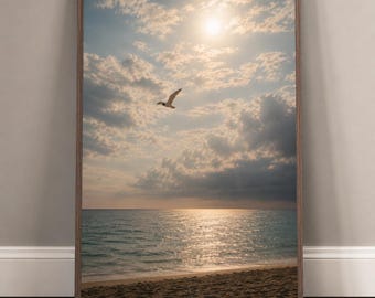 Puede incluir: Una impresión enmarcada de una escena de playa. La imagen presenta un sol brillante brillando a través de las nubes sobre el océano. Una gaviota vuela en el cielo. La playa está en primer plano y el marco es marrón.