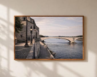 Puede incluir: Una impresión enmarcada de un puente de piedra sobre un río, con un edificio a la izquierda y un cielo azul. El puente tiene un diseño de arco curvo. La impresión está en un marco de madera marrón.