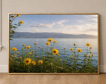 Puede incluir: Una impresión de arte mural de paisaje rectangular en un marco dorado fino que presenta una vista panorámica de un lago azul tranquilo con montañas distantes bajo un cielo suave. En primer plano, girasoles amarillos crecen junto al agua.