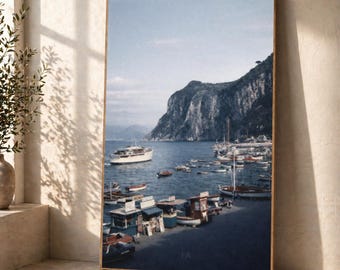 Puede incluir: Una impresión enmarcada de una escena costera con un puerto, barcos, una montaña y un cielo azul claro. La impresión tiene un marco de madera y se exhibe en una pared de color neutro. La escena evoca una sensación de tranquilidad y viaje.