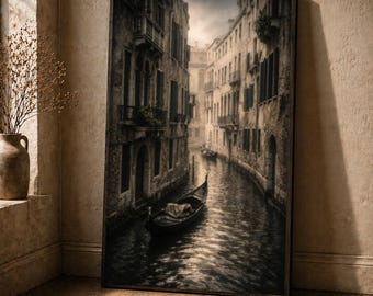 Puede incluir: Una impresión enmarcada representa una escena de canal en Venecia, Italia. La imagen en blanco y negro muestra una góndola en el agua, flanqueada por edificios antiguos. La obra de arte se exhibe sobre un suelo de madera, apoyada contra una pared beige.