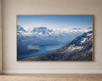 Puede incluir: Una impresión enmarcada de un paisaje de montañas con picos nevados, un lago azul y un cielo despejado. La obra de arte se exhibe en una pared beige sobre un suelo de madera. El marco es de color marrón claro.