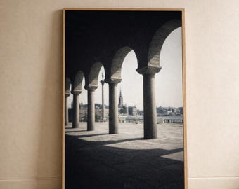 Puede incluir: Impresión enmarcada en blanco y negro que presenta una serie de columnas arqueadas, proyectando sombras sobre un camino de piedra. El fondo revela una ciudad con edificios y una torre. La impresión está en una pared clara.