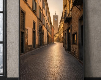 Puede incluir: Una impresión artística enmarcada que muestra una calle estrecha y empedrada en un antiguo pueblo europeo al atardecer. La escena presenta edificios altos de tonos dorados a ambos lados y una torre de reloj de piedra en la distancia.