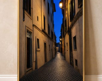 Puede incluir: Una impresión artística enmarcada que muestra un callejón estrecho de adoquines al atardecer. La escena representa edificios altos de color beige con ventanas oscuras que bordean el camino, iluminados por farolas cálidas bajo un cielo azul profundo.