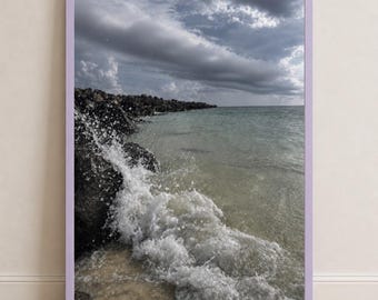 Puede incluir: Una impresión de arte mural enmarcada que muestra una escena costera con rocas oscuras a lo largo de la orilla, olas blancas rompiendo contra las piedras y un cielo nublado sobre el horizonte. La obra está en un marco rectangular de color violeta claro.