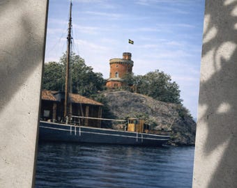 Puede incluir: Impresión de un barco en el agua con una torre en una pequeña isla. El barco es azul oscuro con una cabina de madera. La torre es de color ladrillo con una bandera sueca. El cielo es azul con algunas nubes.