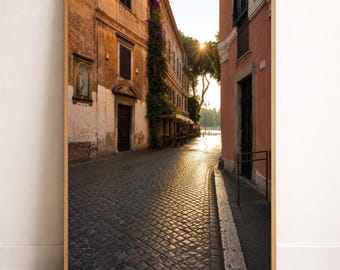 Puede incluir: Una impresión artística enmarcada que representa una estrecha calle empedrada en Roma al atardecer. La escena muestra edificios desgastados de color ocre y melocotón, una enredadera en una pared y un brillante sol al final de la calle.