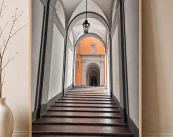Puede incluir: Una impresión de arte mural enmarcada que muestra una vista en perspectiva de una larga escalera de piedra que conduce a través de una serie de pasillos arqueados. La arquitectura está representada en tonos grises, contrastando con un edificio naranja brillante al fondo.