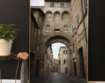 Puede incluir: Una impresión enmarcada de un arco de piedra que conduce a una calle adoquinada. Los edificios son de ladrillo y piedra, con ventanas y detalles arquitectónicos. La impresión está apoyada contra una pared negra, con una planta y un jarrón en primer plano.