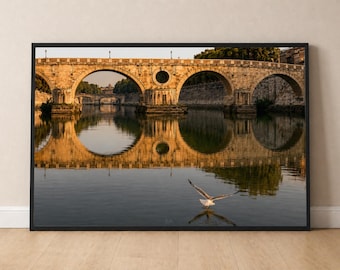 Puede incluir: Una impresión de arte mural enmarcada que muestra un puente de piedra con múltiples arcos sobre un río tranquilo al atardecer. La cálida luz dorada ilumina la mampostería, creando reflejos claros en el agua. Una gaviota vuela sobre el río.