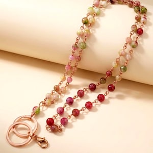 May include: A beaded lanyard with a rose gold chain and hardware. The beads are a mix of colors, including red, pink, green, and white. The lanyard has two key rings and a lobster clasp.