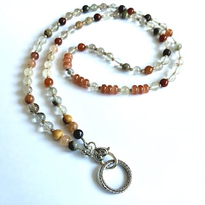 May include: A beaded lanyard featuring a variety of round, translucent, and opaque beads in shades of clear, amber, brown, and black. The strand is finished with a silver-toned heart-shaped clasp attached to a textured circular metal ring.