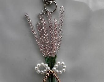 May include: A beaded keychain featuring a lavender bouquet design. The lavender is made of pink beads, tied with a white pearl bow and gold beads. The keychain has a silver clasp.