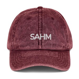 May include: A maroon baseball cap with the embroidered letters "SAHM" in white. The cap has a curved peak and a fabric strap closure. The hat is made of a soft, worn material.