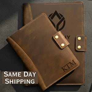 May include: A set of brown leather journals and a pen on a dark surface. The journals have gold-colored button closures and the initials "KTM" are visible on the smaller journal. The larger journal has a logo and the word "NY" printed on it.