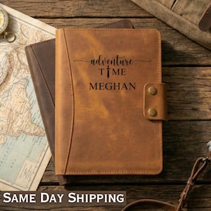 May include: A brown leather journal with the words "adventure time MEGHAN" embossed on the front. The journal has a snap closure and is lying on top of a map and another brown leather journal. A compass and camera are also visible.