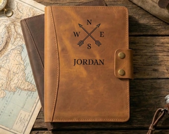 May include: A brown leather journal with the name "JORDAN" engraved below a compass rose design. The journal has a snap closure and is on top of a darker brown leather journal. A map and compass are in the background.