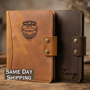May include: Two leather journals, one tan and one dark brown, with brass snap closures. The tan journal features a military-style crest with the name "ROBERT" and "CPT." The dark brown journal is personalized with the name "Terry".