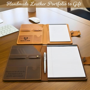 Puede incluir: Dos carteras de cuero abiertas, una beige y otra marr&oacute;n, con blocs de notas rayados. La cartera beige tiene el logotipo de Wayne Enterprises. La cartera marr&oacute;n tiene el texto "Thank you for all you do. Your hard work is greatly appreciated!". El texto "Handmade Leather Portfolio to Gift" est&aacute; en la parte superior.